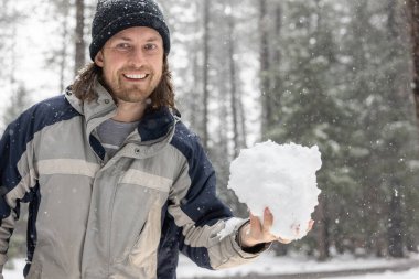 Bir adam elinde bir kartopu tutuyor. Gülümsüyor ve mavi ve gri bir ceket giyiyor. Etrafına kar yağıyor, kış atmosferi yaratıyor.