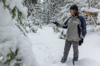 Mavi ceketli ve gri pantolonlu bir adam karda dikilip ağaca bakıyor. Sahne huzurlu ve sakin, karla kaplı zemin ve adamın şapkasıyla