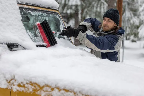 A man is cleaning his car\'s windshield with a snow brush. The scene is set in a snowy environment, and the man is in a good mood