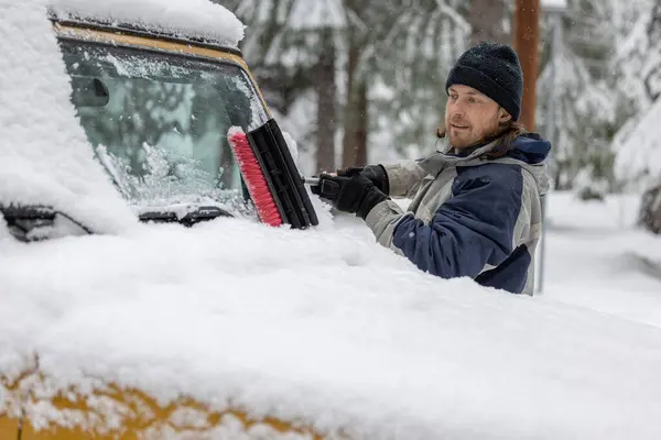 A man is cleaning his car\'s windshield with a snow brush. The scene is set in a snowy environment, and the man is in a good mood