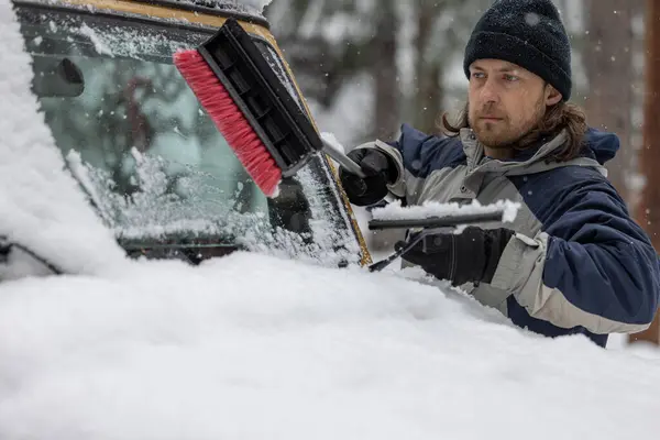 A man is cleaning the windshield of a car with a snow brush. The scene is set in a snowy environment, and the man is focused on his task
