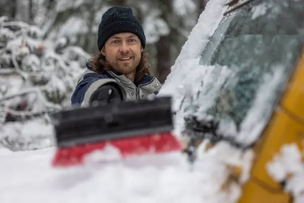 当一个人把雪从汽车上推下来时 他笑了 场景发生在一个大雪的环境中 那个男人戴着一顶黑色的帽子 — 图库照片