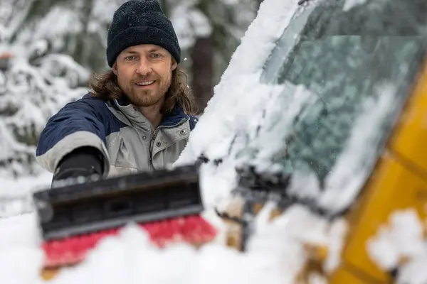 A man is smiling while cleaning his car\'s windshield. The scene is set in a snowy environment, and the man is wearing a black hat
