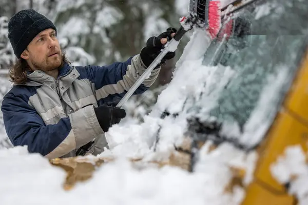 A man in a black hat is clearing snow off a car windshield. The scene is set in a snowy environment, and the man is focused on his task