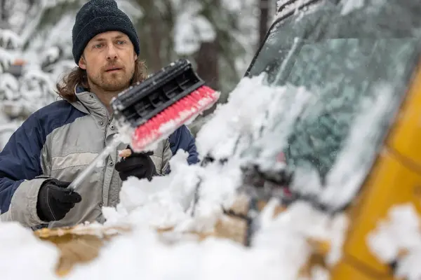 A man is clearing snow off a car with a snow brush. The scene is set in a snowy environment, and the man is focused on his task