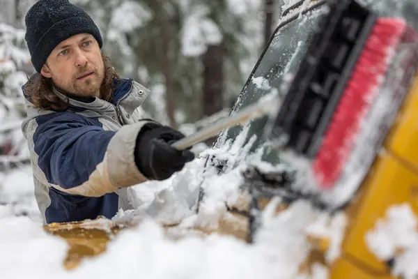A man is cleaning the windshield of a car with a snow brush. The man is wearing a black hat and a blue jacket. The scene is set in a snowy environment