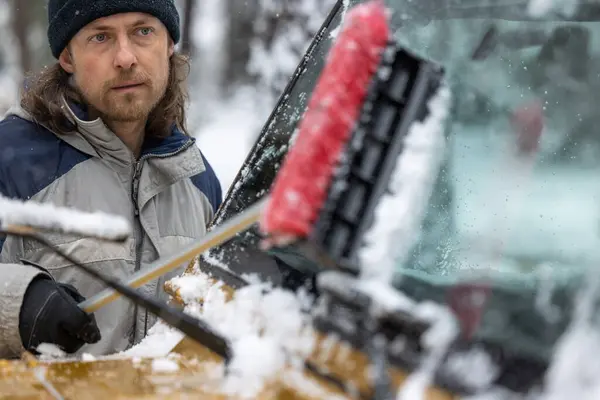 A man is cleaning his car\'s windshield with a snow brush. The scene is set in a snowy environment, and the man is wearing a hat and gloves