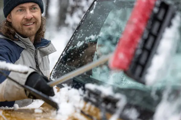 A man is cleaning his car\'s windshield with a snow brush. He is smiling and he is enjoying the task