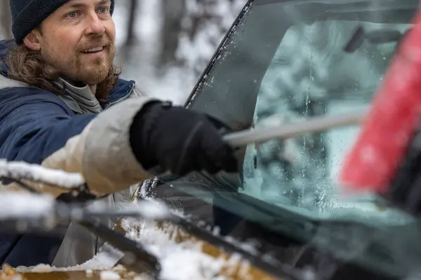 A man is cleaning his car\'s windshield with a snow brush. The man is smiling and he is enjoying the task