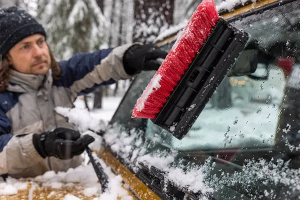 A man is cleaning his car\'s windshield with a brush. The scene is set in the snow, and the man is wearing a hat