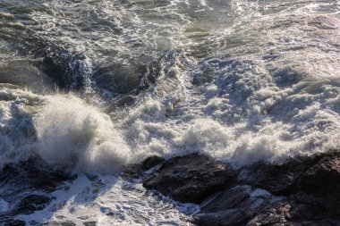 Büyük bir dalga kayalık bir sahile çarpıyor. Su beyaz ve köpüklü ve kayalar sahnenin her tarafına saçılmış. Sahne güçlü ve dramatik.