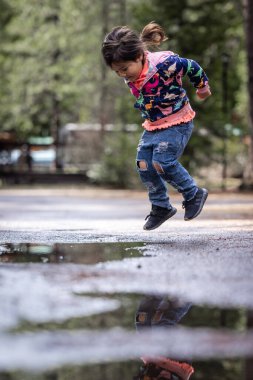 Genç bir kız su birikintisine atlıyor. Neşe ve neşe kavramı, kız suya su sıçratırken eğlenirken. Su birikintisi çevreyi yansıtıyor.