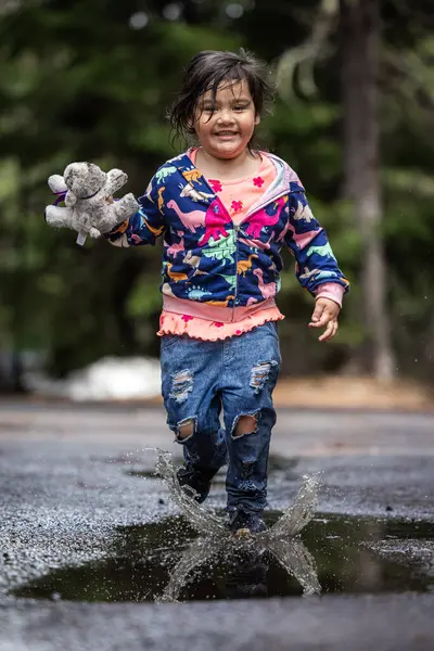 A young girl is playing in the rain, holding a stuffed animal. The scene is playful and joyful, with the girl enjoying the moment and the rain