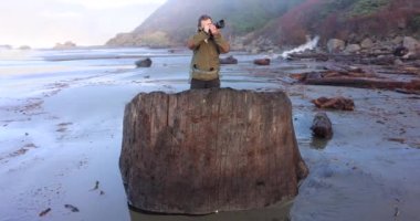 Bir adam okyanusta bir kütüğün üzerinde duruyor. Sahilin fotoğrafını çekiyor..