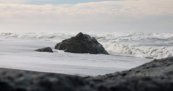 大きな岩が海の隣のビーチに座っています 岩は水に囲まれ 波はそれに衝突しています シーンは穏やかで平和です — ストック動画