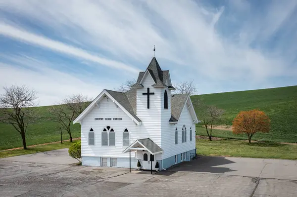 一个顶部有十字架的白色小教堂 教堂四周长满了树和草 天空是蓝色的 背景上有云彩 — 图库照片