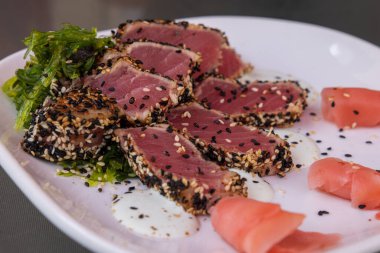 Üzerinde ince dilimlenmiş ton balığı ve susam tohumları olan bir tabak yemek. Tabak beyaz ve yemekler görsel olarak çekici bir şekilde düzenlenmiş.