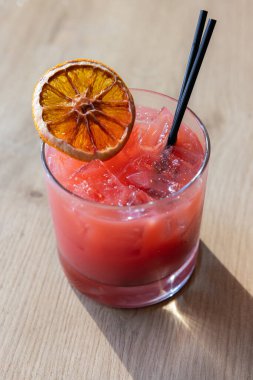Üstünde bir dilim portakal olan pembe bir bardak içki. İçki kısa bir bardakta, siyah bir pipetle servis edilir.
