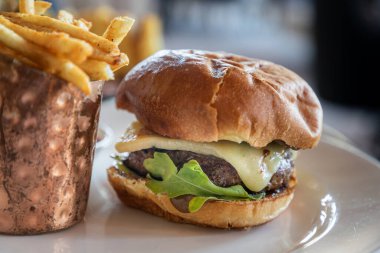 Marul ve peynirli sığır filetosu hamburgeri, metal bir kasenin yanında patates kızartmasının yanında beyaz bir tabakta oturur. Hamburger görüntünün ana odak noktasıdır ve kase biraz renk katan dekoratif bir elementtir.