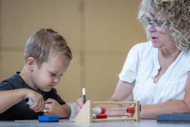 Genç bir çocuk matematik probleminde ona yardım eden bir öğretmenle aynı masada oturuyor. Kadın gözlük takıyor ve yüzünde ciddi bir ifade var. Çocuk elimizdeki işe odaklanmış durumda.