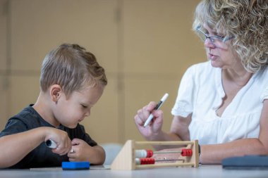 Genç bir çocuk matematik probleminde ona yardım eden bir öğretmenle aynı masada oturuyor. Kadın gözlük takıyor ve yüzünde ciddi bir ifade var. Çocuk elimizdeki işe odaklanmış durumda.