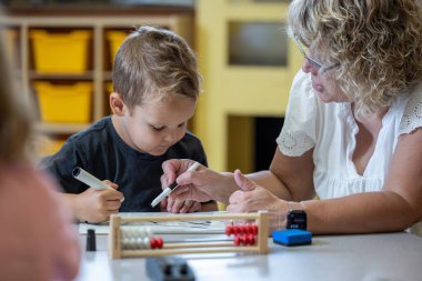 Genç bir çocuk matematik probleminde ona yardım eden bir öğretmenle aynı masada oturuyor. Kadın gözlük takıyor ve yüzünde ciddi bir ifade var. Çocuk elimizdeki işe odaklanmış durumda.