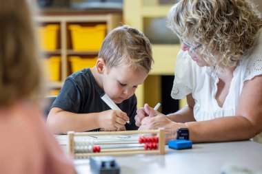Genç bir çocuk matematik probleminde ona yardım eden bir öğretmenle aynı masada oturuyor. Kadın gözlük takıyor ve yüzünde ciddi bir ifade var. Çocuk elimizdeki işe odaklanmış durumda.