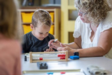 Genç bir çocuk matematik probleminde ona yardım eden bir öğretmenle aynı masada oturuyor. Kadın gözlük takıyor ve yüzünde ciddi bir ifade var. Çocuk elimizdeki işe odaklanmış durumda.