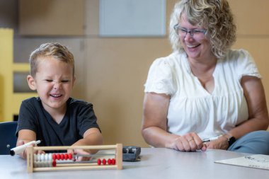 Genç bir çocuk bir abaküs ile masada oturuyor ve sorunu çözerken öğretmeniyle birlikte gülüyor..