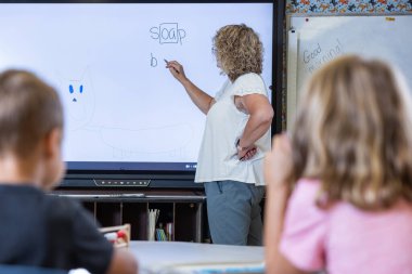 Bir öğretmen üzerinde sabun yazan bir beyaz tahtanın önünde duruyor. Bir grup öğrenci kurulun önünde oturuyor, öğretmeni izliyorlar.