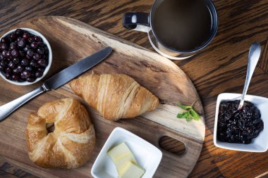 Bir tabak kruvasan ve bir fincan kahve tahta bir kesme tahtasının üzerinde oturur. Kruvasanlar taze böğürtlen reçeli ile yenmeye hazır gibi görünüyorlar..