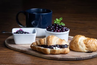 Yaban mersini reçelli bir tabak kruvasan ve bir fincan kahve ahşap bir masada oturur. Kruvasanlar ahşap bir tepside ve kahve de mavi bir kupada. Rahatlık ve rahatlama kavramı