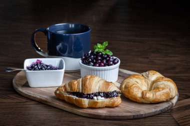 Yaban mersini reçelli bir tabak kruvasan ve bir fincan kahve ahşap bir masada oturur. Kruvasanlar ahşap bir tepside ve kahve de mavi bir kupada. Rahatlık ve rahatlama kavramı