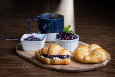 Yaban mersini reçelli bir tabak kruvasan ve bir fincan kahve ahşap bir masada oturur. Kruvasanlar ahşap bir tepside ve kahve de mavi bir kupada. Rahatlık ve rahatlama kavramı