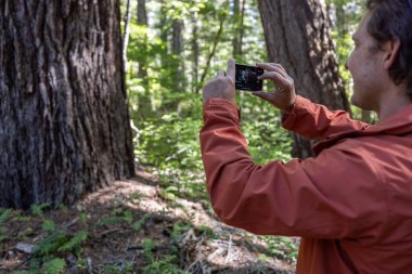 Bir adam kameralı telefonuyla bir ağacın fotoğrafını çekiyor. Adam turuncu bir ceket giyiyor..