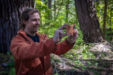 Bir adam kameralı telefonuyla bir ağacın fotoğrafını çekiyor. Adam turuncu bir ceket giyiyor..