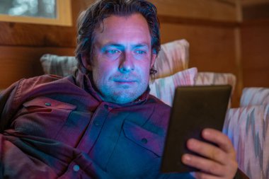 Bir adam kanepeye oturmuş e-kitap okuyor. Gülümsüyor ve eğleniyor.