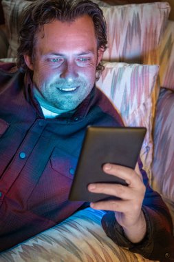 Bir adam kanepeye oturmuş e-kitap okuyor. Gülümsüyor ve eğleniyor.