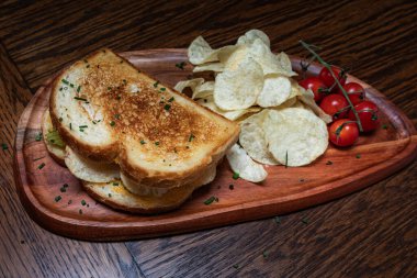 Bir tabakta patates cipsi ve yanında domates olan bir sandviç. Sandviç ahşap bir tabakta servis edilir..