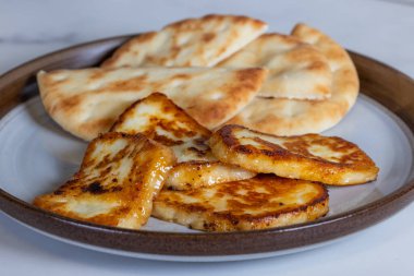 Bir tabak kızarmış halloumi peyniri ve yanında da pita ekmeği. Yemekler altın sarısı ve lezzetli görünüyor..