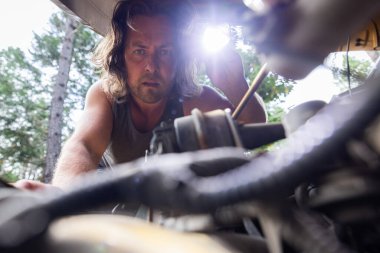 Adam dışarıdaki ağaçlık alanda el feneri kullanarak araba motorunu inceliyor. Doğal ışık altında el yapımı otomobil tamiri, kaputun altından alındı..