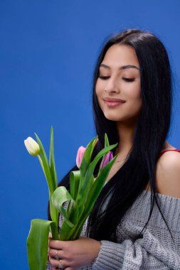 Elinde lalelerle stüdyoda mavi arka planda poz veren güzel bir genç kadın.