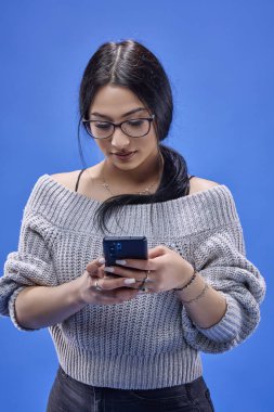 Stüdyoda mavi bir arka planda elinde akıllı bir telefonla poz veren güzel, genç bir esmer..