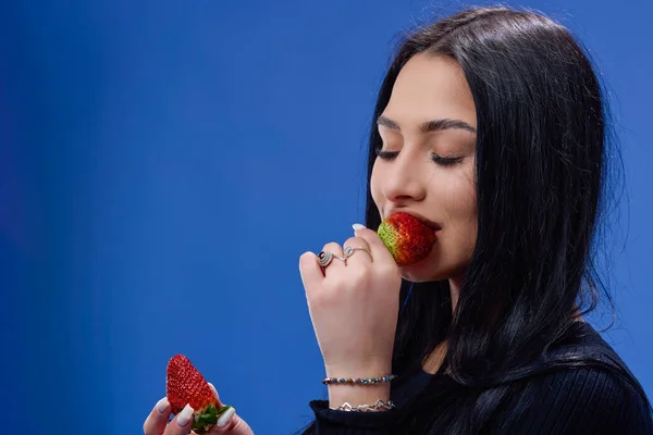 一个年轻美丽的黑发姑娘吃草莓的画像 背景是蓝色的 — 图库照片