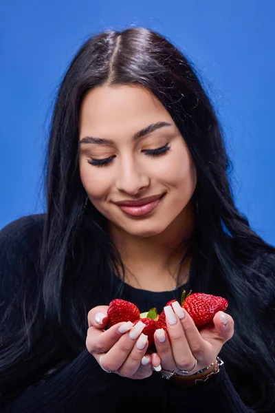一个年轻美丽的黑发姑娘吃草莓的画像 背景是蓝色的 — 图库照片