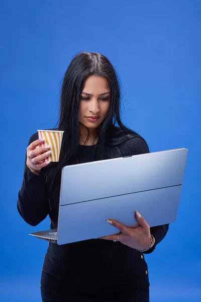 一个年轻貌美的黑发姑娘站在演播室里 蓝色背景下 拿着一杯水和一台笔记本电脑的画像 — 图库照片