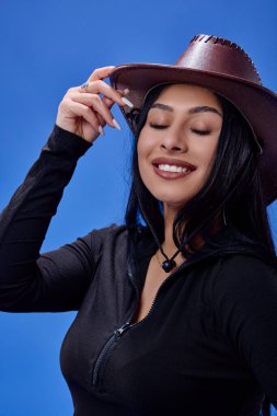 Stüdyoda poz veren uzun saçlı ve şapkalı genç bir esmer, mavi arka planda izole edilmiş..