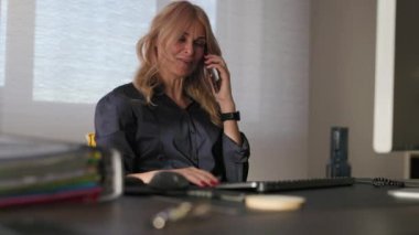 İş kadını ofiste bilgisayarın başına oturur ve telefonla konuşur.