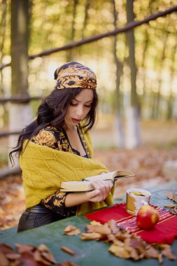Kafasında eşarp ve battaniyesi olan güzel esmer, sonbahar manzarasında bir kitap, bir fincan ve bir elmayla bir masada oturuyor..