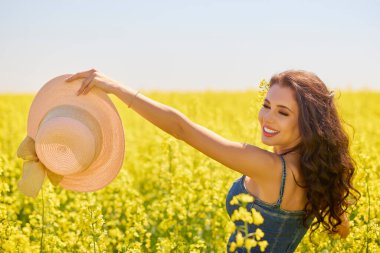 Güneşli bir günde çiçek açan kolza tarlasında poz veren şapkalı güzel bir kadın.